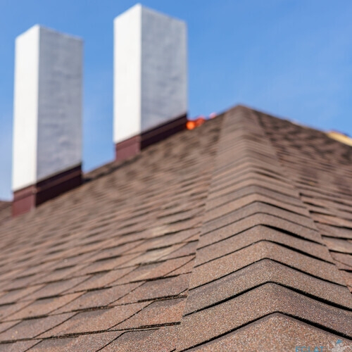 Vertical photo of asphalt tile roof with two white and concrete chimney on new home under construction against blue sky on background